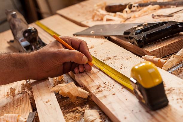 Close-up of a hand measuring a wooden board with a yellow tape measure in a woodworking workshop.