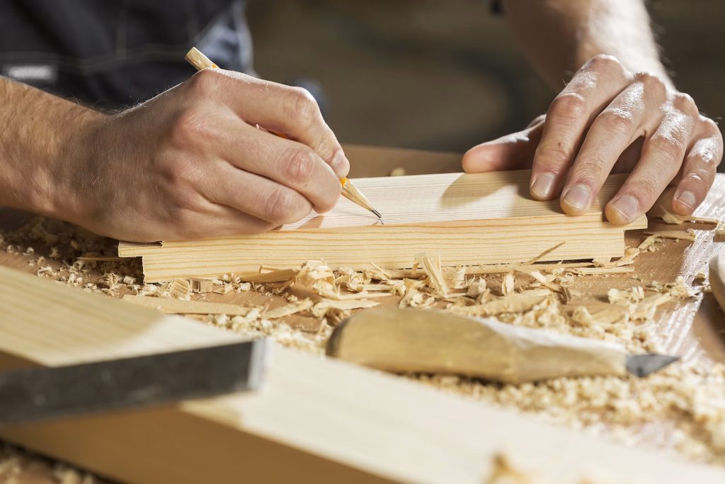 Close-up of hands shaping a wooden plank with a carving tool, wood shavings scattered around.